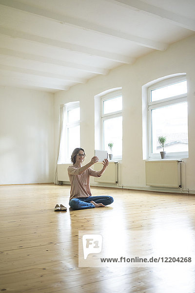Lächelnde reife Frau sitzt auf dem Boden in einem leeren Raum und nimmt Selfie mit Tablette.