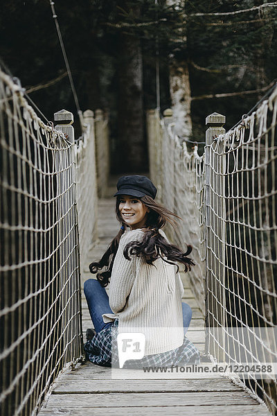 Porträt einer lächelnden jungen Frau  die auf einer Hängebrücke sitzt.