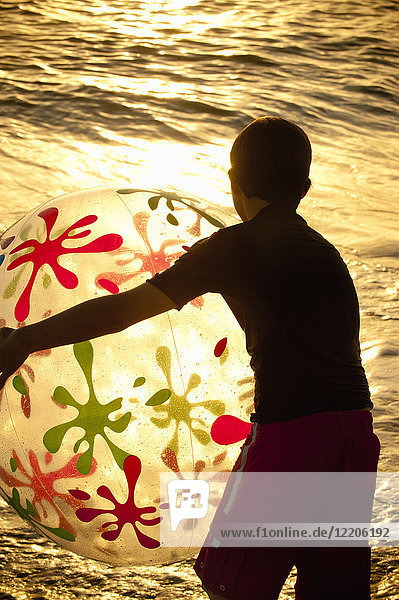 Silhouette eines kaukasischen Jungen  der bei Sonnenuntergang einen Strandball hält