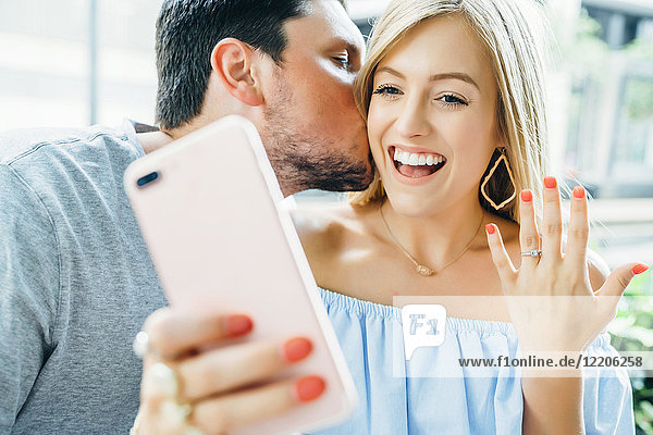 Caucasian couple holding hands and posing for selfie with engagement ring