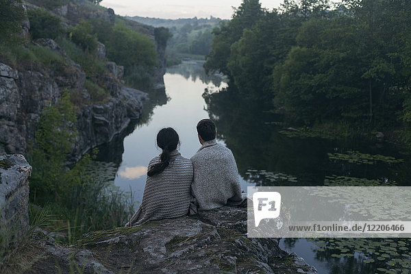 Caucasian couple wrapped in blanket sitting on rock admiring river