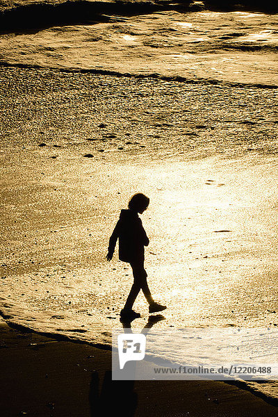 Silhouette eines gemischtrassigen Jungen  der bei Sonnenuntergang am Strand spazieren geht
