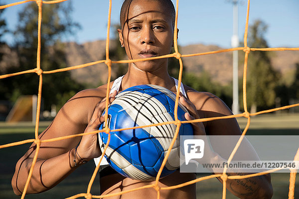 Porträt einer Frau hinter einem Ziegennetz  die einen Fussball hält und in die Kamera schaut