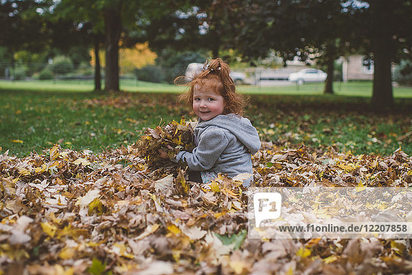 Portrait of red haired female toddler in park with bundles of autumn leaves