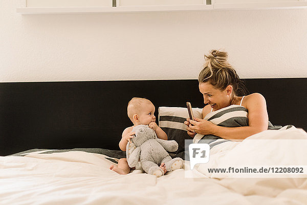 Mutter fotografiert Baby-Tochter im Bett sitzend mit Plüschtier