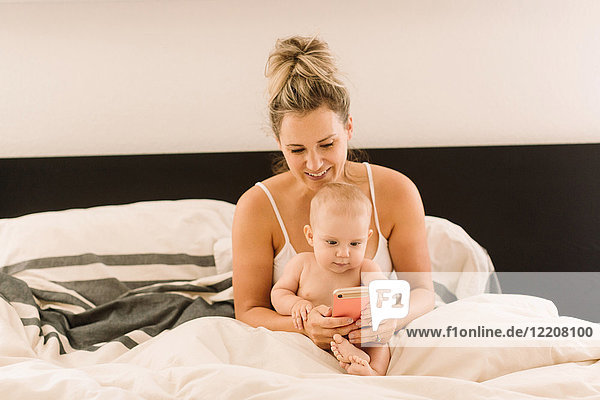 Baby girl and mother sitting up in bed looking at smartphone