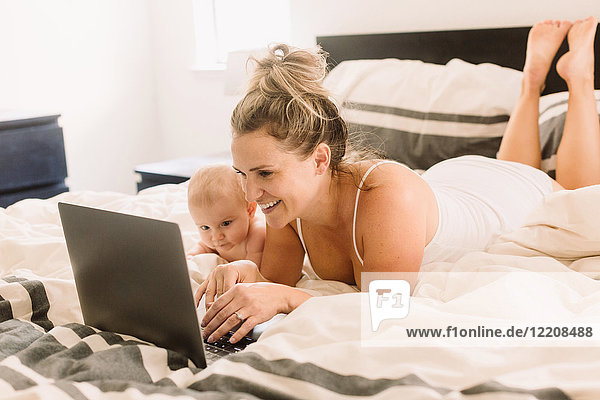 Baby girl and mother lying on bed looking at laptop