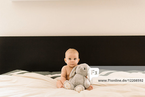 Portrait of cute baby girl sitting up on bed with soft toy