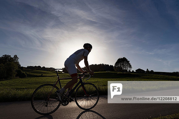 Hintergrundbeleuchtete Ansicht eines jungen männlichen Radfahrers  der bei Sonnenuntergang die Landstraße hinauffährt