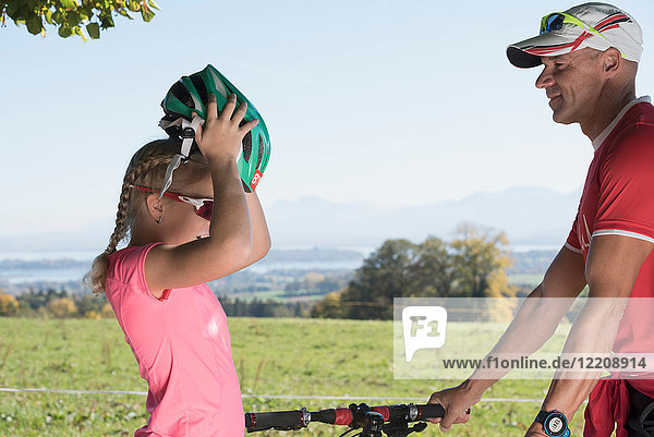 Father watching girl cyclist putting on bicycle helmet