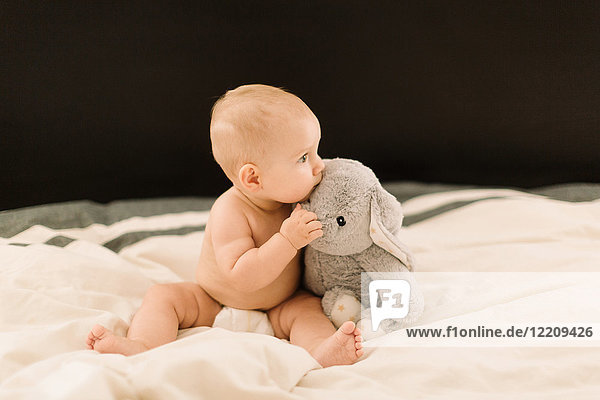 Cute baby girl sitting up on bed with soft toy