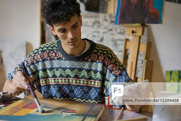 Männlicher Künstler schaut auf Smartphone  während er im Künstleratelier Leinwand malt