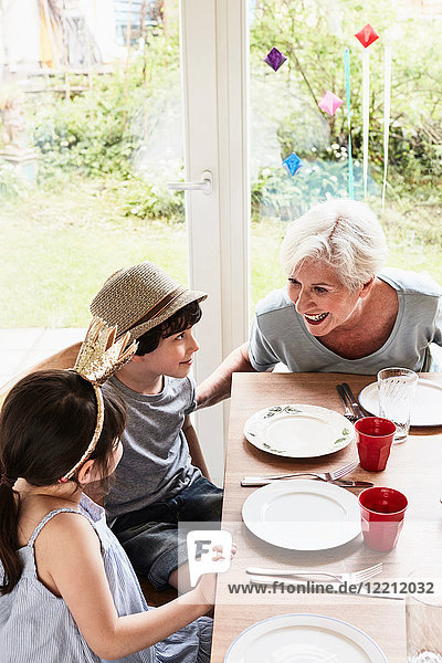 Großmutter sitzt mit Enkel und Enkelin am Küchentisch und lächelt
