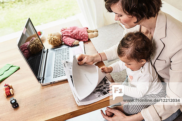 Mutter sitzt mit ihrer kleinen Tochter  schaut Papierkram durch  Laptop auf dem Tisch vor ihr