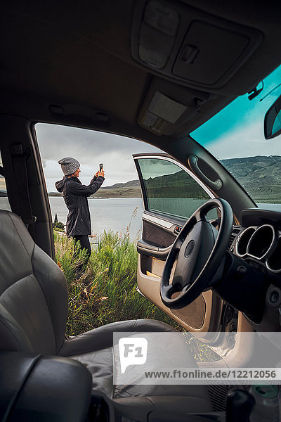 Junge Frau steht neben dem Dillon-Stausee  hält ein Smartphone in der Hand  Blick durch ein geparktes Auto  Silverthorne  Colorado  USA