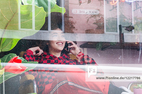 Young woman sitting indoors  using smartphone  viewed through window