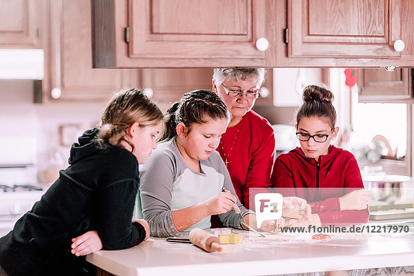 Drei Mädchen und Großmutter mit Keksteig auf Spachtel an der Küchentheke