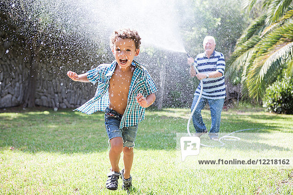 Boy running away from grandfather spraying hosepipe in garden