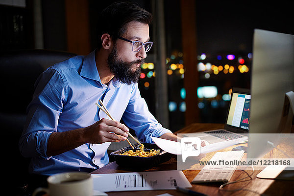 Junger Geschäftsmann betrachtet Computer und isst abends am Büroschreibtisch zum Mitnehmen