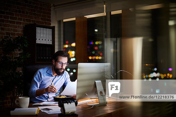 Young businessman reading paperwork and eating takeaway at office desk at night