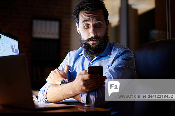 Junger Geschäftsmann schaut nachts am Büroschreibtisch auf Smartphone