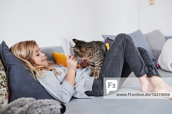 Young girl lying on sofa  reading book  pet cat peering round book