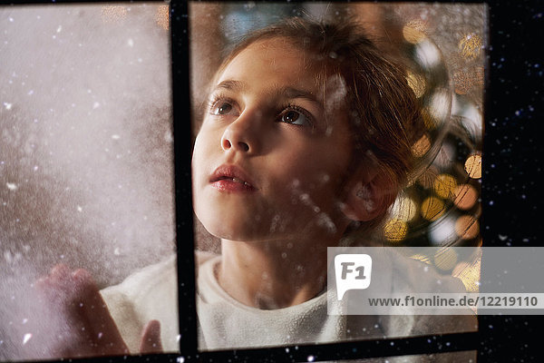 Junges Mädchen schaut aus dem Fenster  im Hintergrund hinter ihr ein Weihnachtsbaum  durchs Fenster betrachtet
