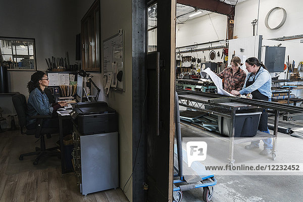 Zwei Frauen an einem Schreibtisch im Bürobereich einer Metallwerkstatt  Frau steht an einer mobilen Werkbank.