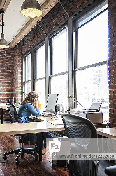 Caucasian woman at office workstation near a large bank of windows.