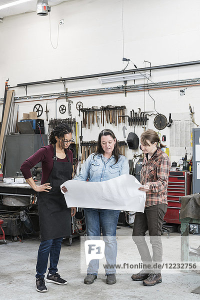 Drei Frauen stehen in der Metallwerkstatt und halten einen technischen Bauplan in der Hand.