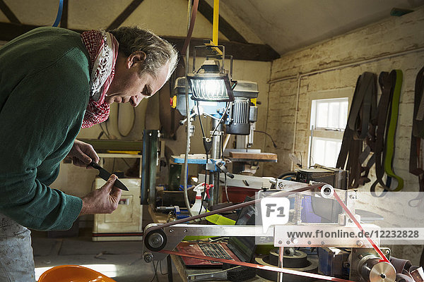 Ein Handwerker betrachtet eine kleine Metallform  ein Küchenmesser  das sich darauf vorbereitet  den Griffabschnitt auf einer rotierenden Schleifscheibe zu glätten.