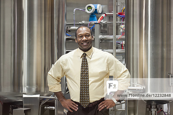African American male management person in a shirt and tie standing in front of processing tanks in a bottling plant.