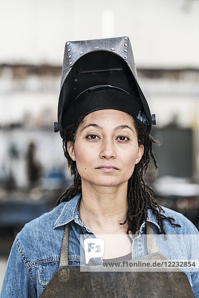 Porträt einer Frau mit Schürze und Schweißermaske  die in einer Metallwerkstatt steht und in die Kamera schaut.