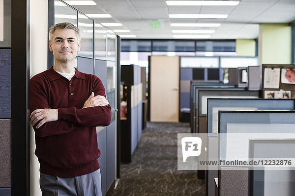 A casually dressed Caucasian businessman standing next to his office in a room full of cubicles.