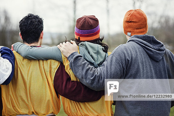 View from behind of three friends with their arms around one another in sports kit and woolly hats.