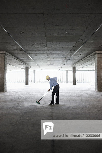Asian businessman cleaning up with a broom in a large empty raw office space.