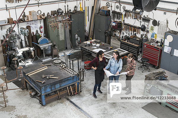 Hochwinkelaufnahme von drei Frauen  die in einer Metallwerkstatt stehen und eine technische Blaupause halten.