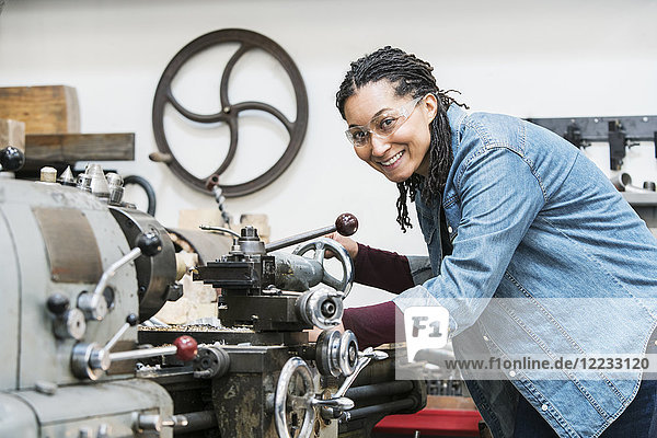 Lächelnde Frau mit Schutzbrille  die in einer Metallwerkstatt an einer Maschine steht und in die Kamera schaut.
