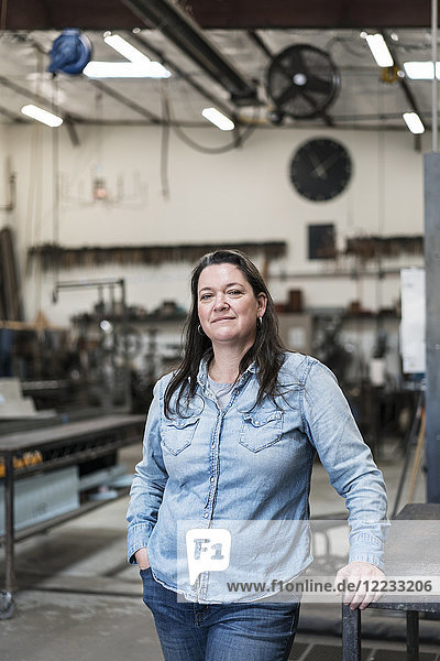 Frau mit braunen Haaren in Denim-Hemd steht in Metallwerkstatt und lächelt in die Kamera.