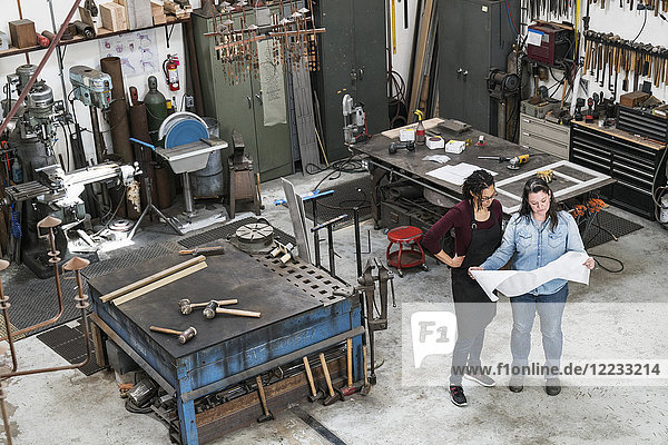 High angle view of two women standing in metal workshop  holding technical blueprint.