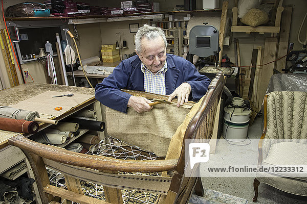 Kaukasischer älterer männlicher Tapezierer  der in seiner Autowerkstatt an einem Stuhl arbeitet.