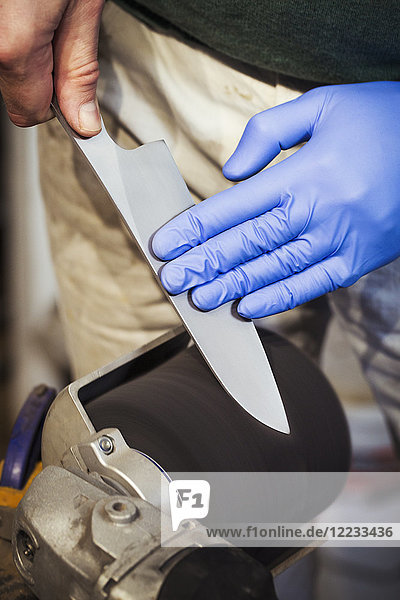 Nahaufnahme eines Handwerkers in einer Werkstatt  der die Klinge eines Küchenmessers mit einer rotierenden Schleifscheibe schärft und schleift.
