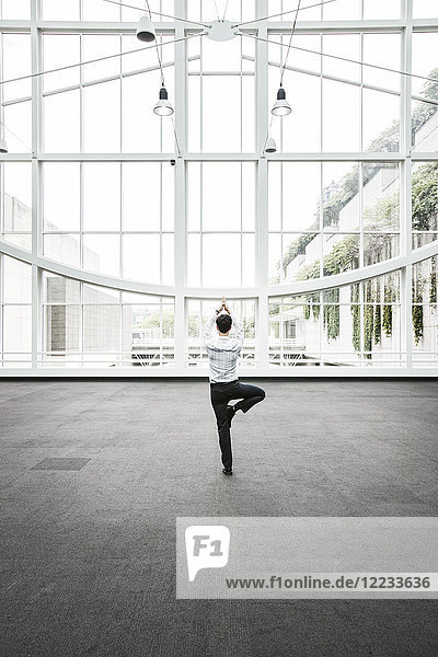 Geschäftsmann entspannt sich bei einer Yoga-Pose in einem großen  mit Glas überdachten Gang.