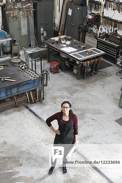Hochwinkelaufnahme einer Frau mit Brille und Schürze  die in einer Metallwerkstatt steht und in die Kamera schaut.