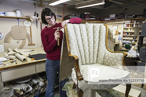 Eine junge Tapeziererin mit einem Reißnagelhammer auf einem Stuhl in einer Tapezierwerkstatt.