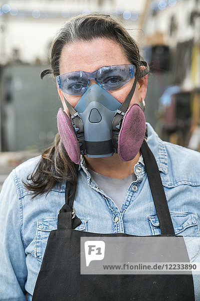 Porträt einer Frau mit Schutzbrille und Staubmaske  die in einer Metallwerkstatt steht und in die Kamera schaut.