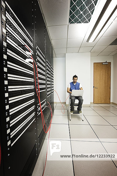 Computertechniker  der an einem Lap-Top-Computer arbeitet  während er diagnostische Arbeiten an einem Server in einem größeren Computer-Serverraum durchführt.