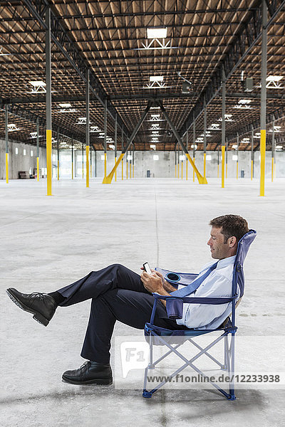 Caucasian man sitting in the middle of a new empty warehouse and using cell phone.