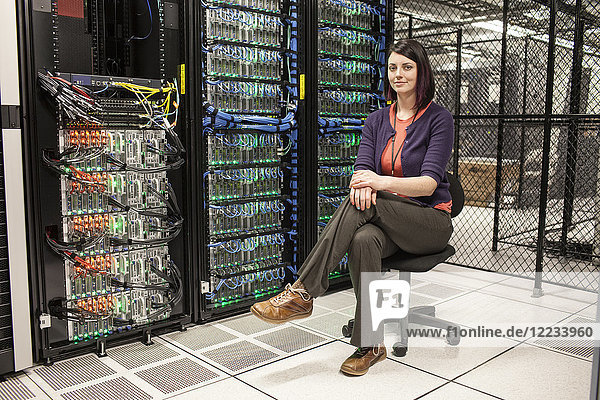 Caucasian woman technician sitting in an aisle of a large computer server farm.