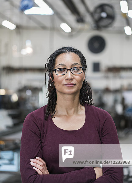 Woman with brown hair wearing glasses standing in metal workshop  smiling at camera.